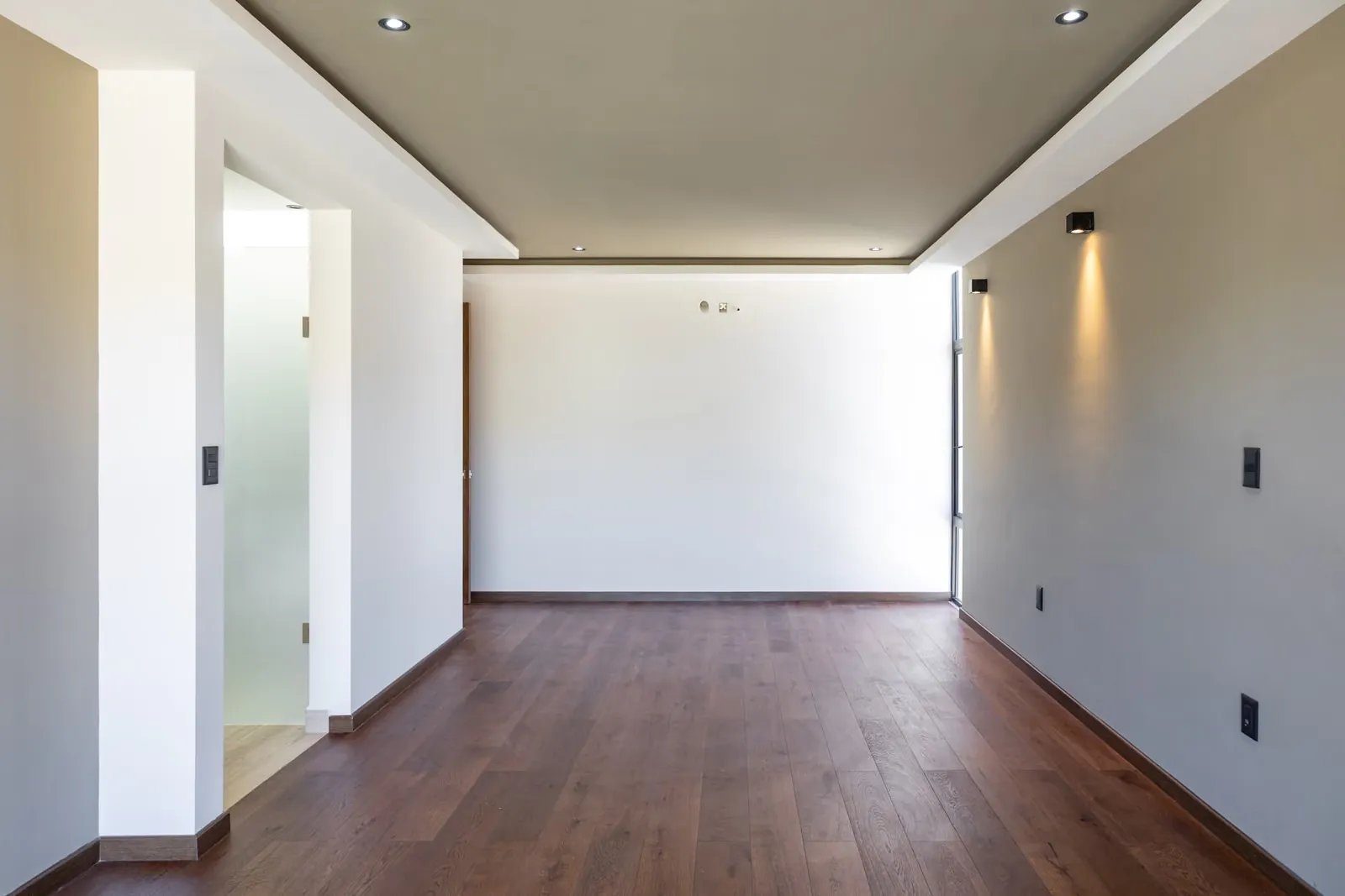 Habitación vacía de Casa Roqueño 68 con piso de tablones en tono nogal, muros blancos y gris claro, arbotantes en el muro y luces empotradas en el plafón