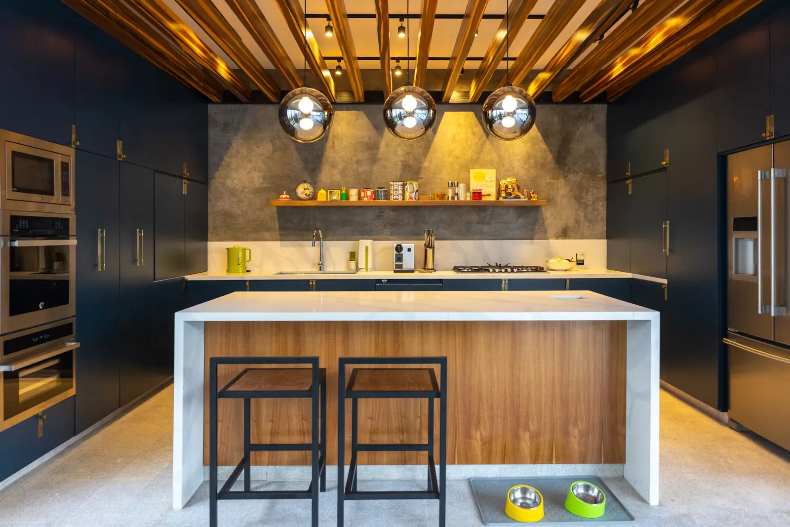 Cocina con gabinetes azul marino de piso a techo, isla blanca con frente de madera y tres lámparas colgantes bajo un plafón de listones de madera